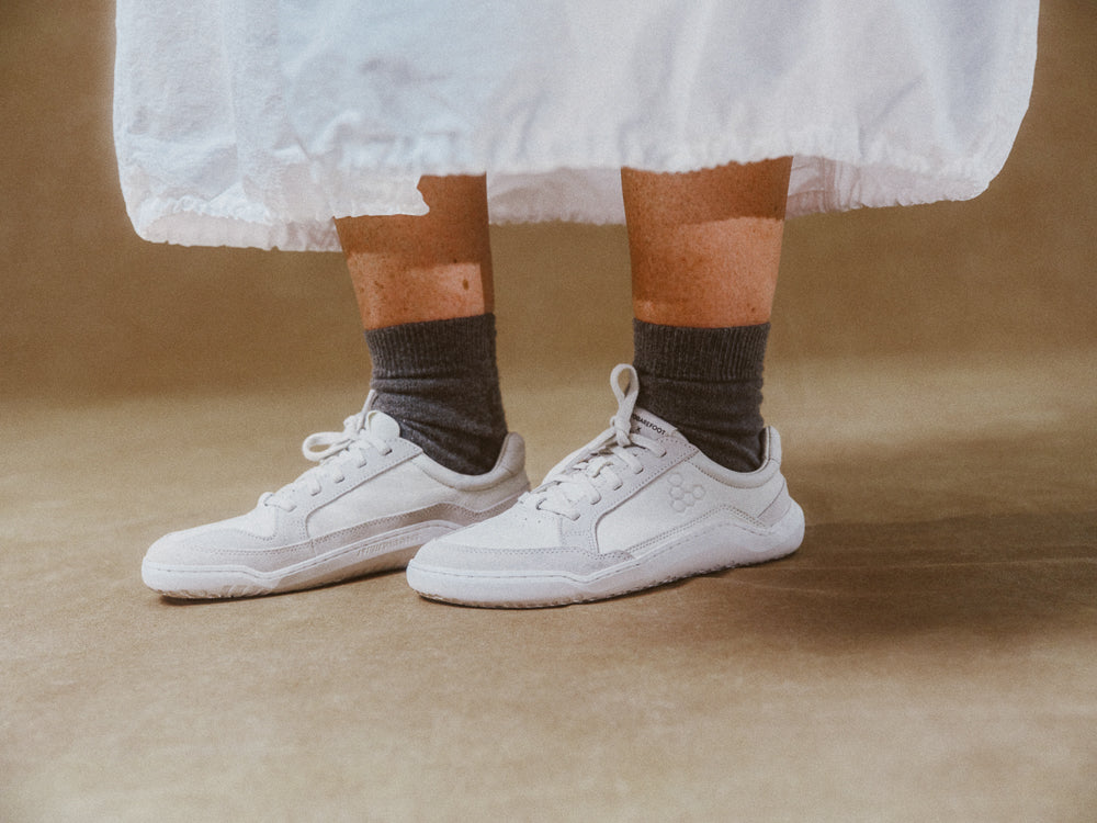White sneakers worn with brown socks on a brown background wearing Limestone Gobi Sneaker II Premium Leather by Vivobarefoot