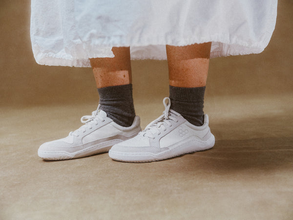 White sneakers worn with brown socks on a brown background wearing Limestone Gobi Sneaker II Premium Leather by Vivobarefoot